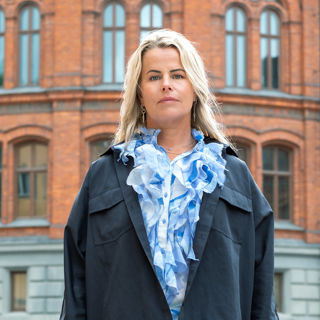 Anna Olskog med neutral min, stående utomhus iklädd mörkblå jacka och en ljusblå blus med en massa krås/volanger.